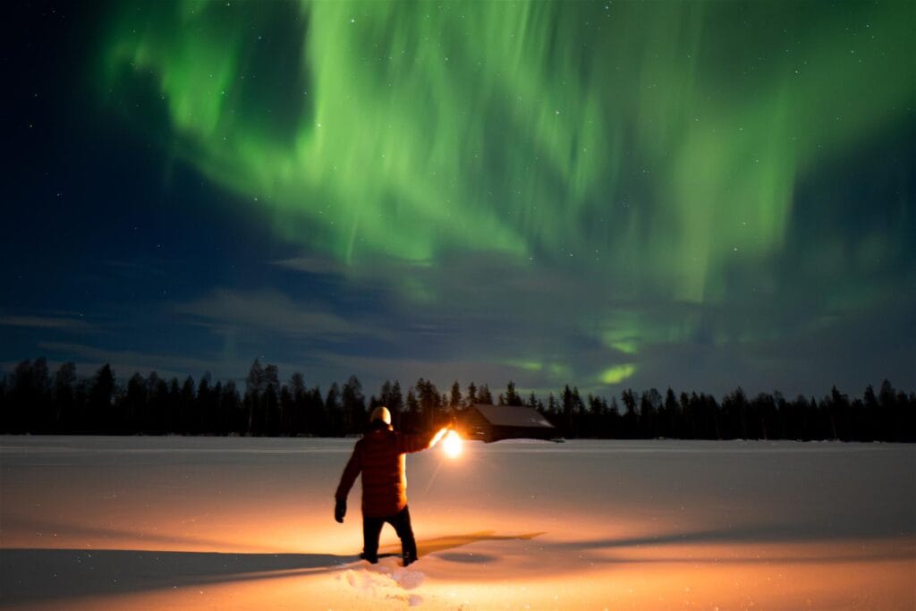 Mies kahlaa lumessa lyhty kädessä. Pimeällä taivaalla näkyy revontulia,