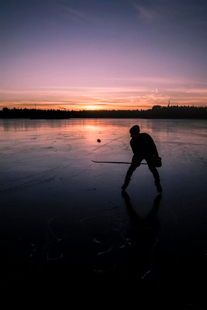 Mies luistelee järven jäällä ja pompottaa jääkiekkoa mailallaan. Aurinko laskee.