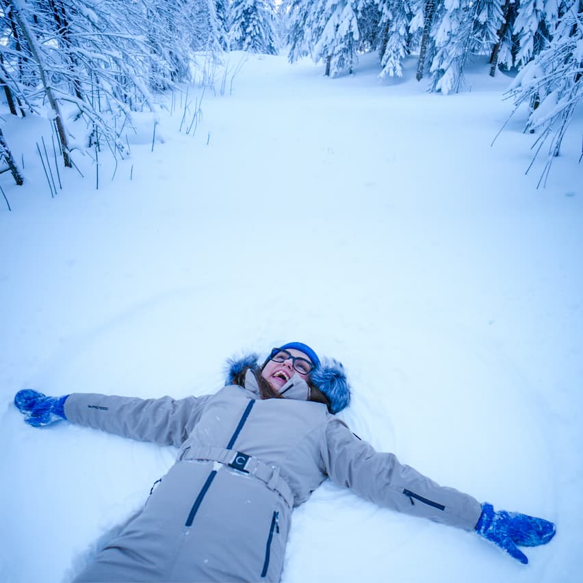 Nainen beigessä haalarissa tekemässä lumienkeliä hankeen.