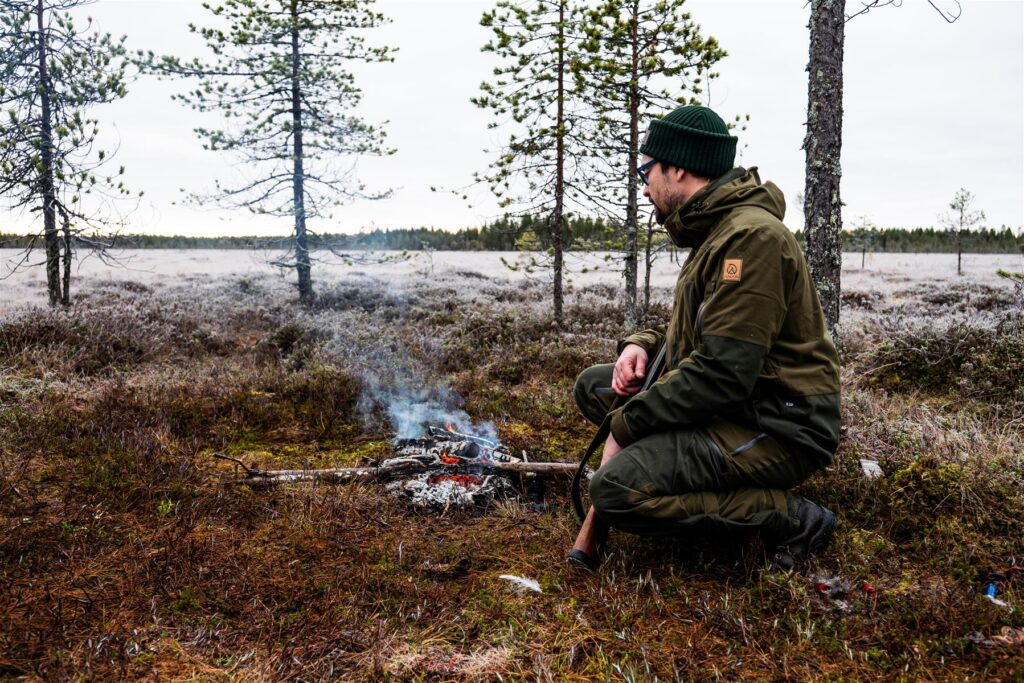 Mies istuu suon syksyllä suon laidalla. Edessä on nuotio ja miehen kädessä on haulikko.