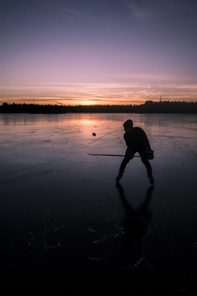 Mies luistelee järven jäällä ja pompottaa jääkiekkoa mailallaan. Aurinko laskee.